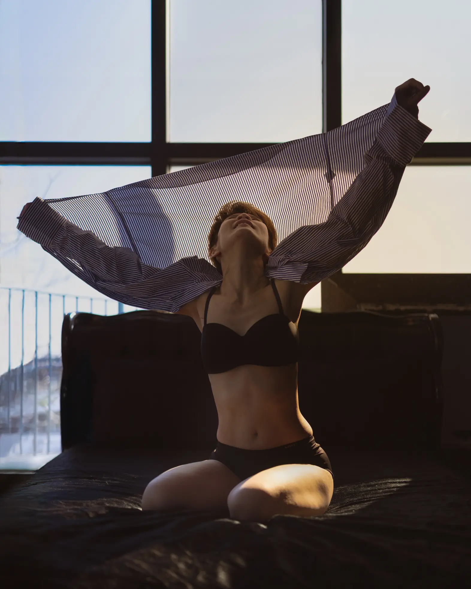 Photograph of a woman sitting on a bed with arms raised up in a gesture of release